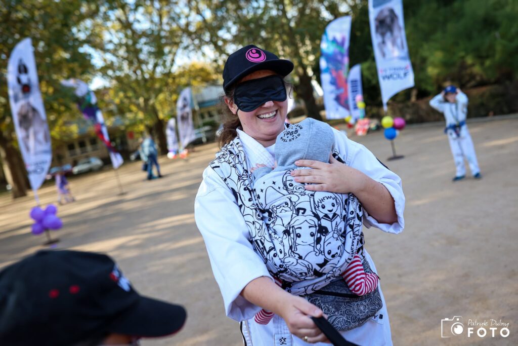 Uczestniczka Biegu w Ciemno z czarną opaską zasłaniającą oczy niesie dziecko w chuście podczas wydarzenia na Jasnych Błoniach w Szczecinie.