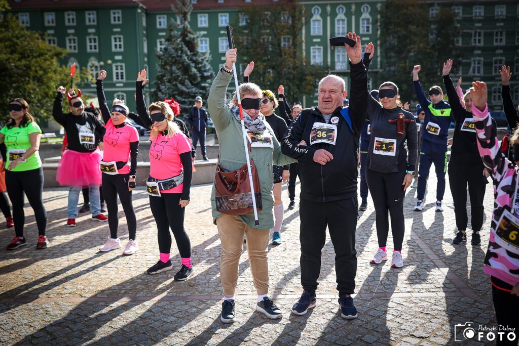 Zdjęcie z jednej z poprzednich edycji Biegu w Ciemno. Na placu przed charakterystyczną zieloną zabudową stoi grupa uczestników wydarzenia. Wiele osób ma czarne opaski zasłaniające oczy i unosi jedną rękę do góry. W centrum znajduje się kobieta w jasnej kurtce, z oczami zasłoniętymi czarną opaską, trzymająca w uniesionej dłoni białą laskę. Obok niej stoi uśmiechnięty mężczyzna z numerem startowym 36, również unoszący rękę. W tle widać kolejnych uczestników w sportowych, kolorowych strojach. Zdjęcie pokazuje wspólne doświadczanie wydarzenia przez osoby z różnymi doświadczeniami i sposobami poruszania się. Autorem fotografii jest Patryk Dulny FOTO.